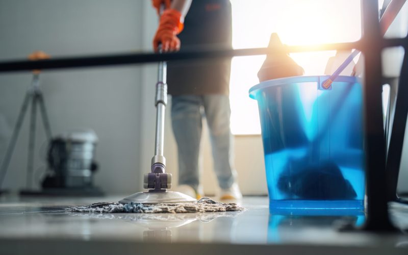 A cleaning woman is standing inside a building holding with a blue tank on the side facilities for tidying up in her hand.