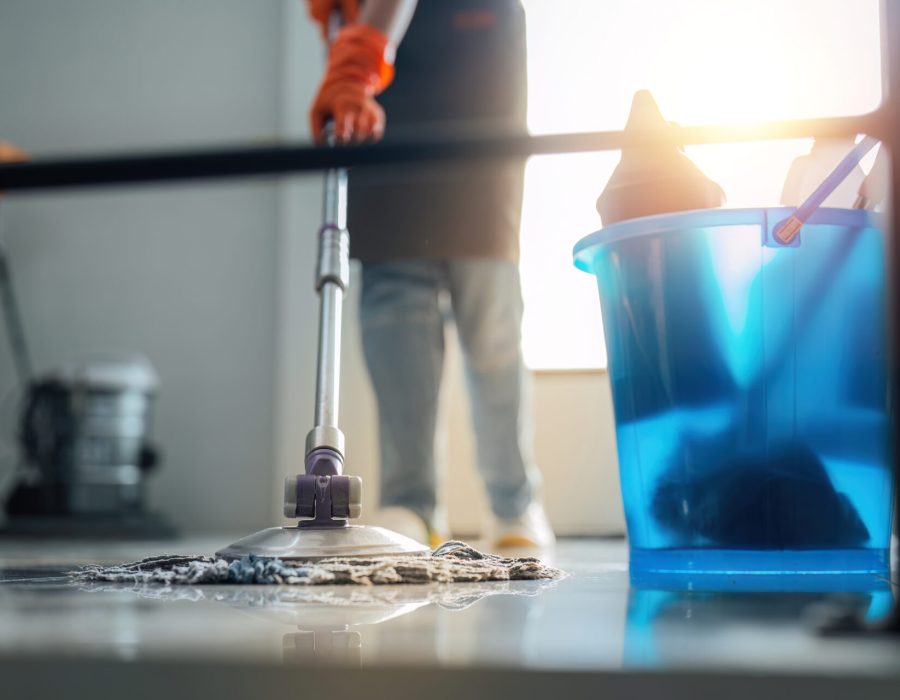 A cleaning woman is standing inside a building holding with a blue tank on the side facilities for tidying up in her hand.